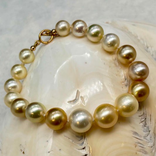 A strand bracelet made of large, lustrous freshwater pearls in champagne, cream, and soft gold tones, finished with a gold clasp, displayed on a white seashell.