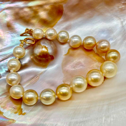 A strand of large, round freshwater-style pearls with a warm cream to pale gold luster, threaded on a cord and displayed on a glossy, iridescent mother-of-pearl shell. A gold clasp is visible on one end.